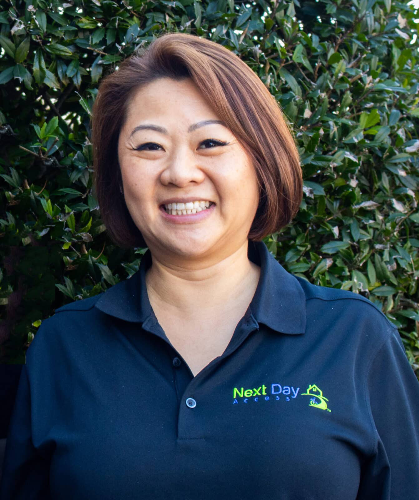 Brown-haired woman smiling in a black "Next Day Access" polo shirt against a green hedge.