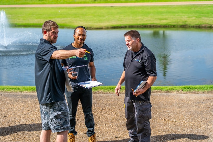 Three men in uniform stand outside next to a pond and discuss build plans