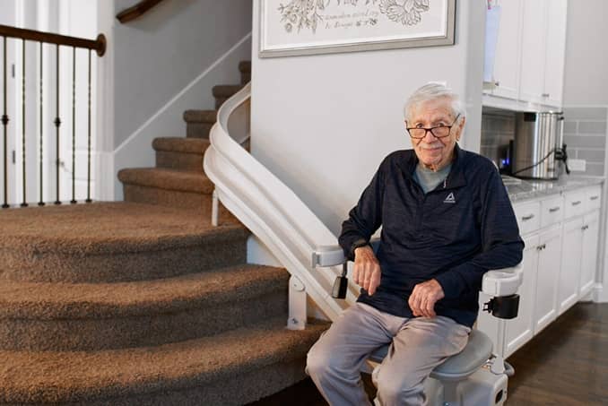 an elderly man sits at the bottom of his home stairs on a chair lift