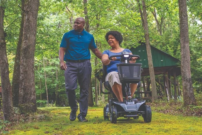 an elderly man walks next to a woman in a motorized scooter on a hiking trail