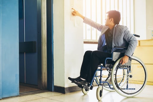 a well dressed man in a wheelchair hits the button to an elevator in an office building