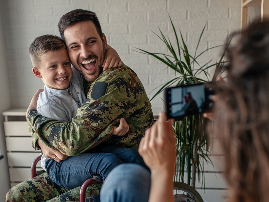 disabled veteran in uniform hugs onto his son while the boys mother takes a photo
