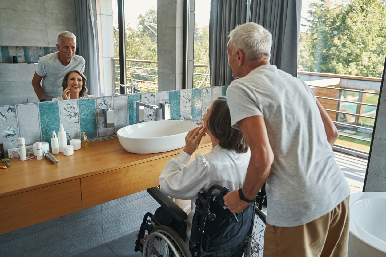 Elderly Caucasian woman with disability checking her face in mirror while smiling man holding her wheelchair