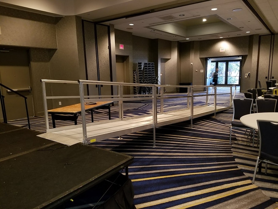 A hotel business center with blue and gold carpet contains a black stage with a metal wheelchair ramp attached