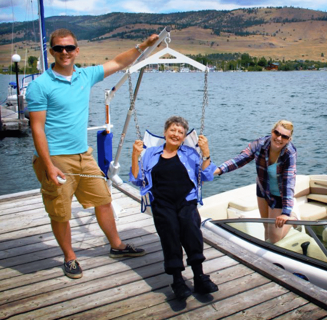 a group assists a woman in a pool lift on a boat dock and lowers her into a boat