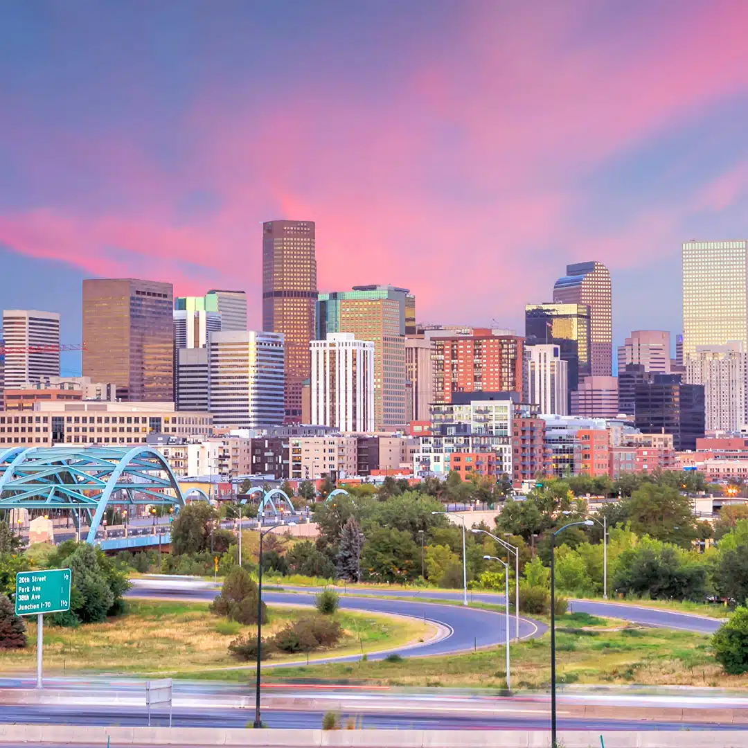 Denver skyline at sunset with pink and purple clouds above the downtown buildings and a highway in the foreground.